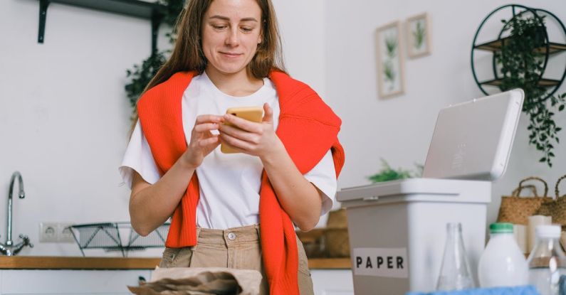 Compost Bin - Cheerful female browsing mobile phone and standing at table with sorted paper and plastic waste